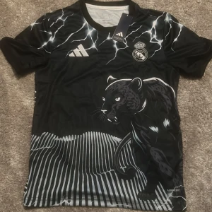 Svart fotbollströja med pantermotiv från Adidas - Cool svart Real Madrid fotbollströja från Adidas med ett unikt pantermotiv och blixtdetaljer. Har aldrig används och storleken är medlens xs och S