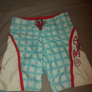 Rutig badshorts från Billabong - Snygga badshorts från Björn Borg med blått och vitt rutmönster och röda detaljer. Shortsen har snörning framtill och en praktisk ficka på sidan. Perfekta för en dag på stranden eller vid poolen. Goofy ai😭