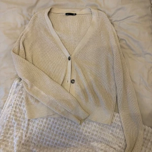 Beige stickad kofta från 157 - Säljer en stilren beige stickad kofta från 157. Koftan har en v-ringad design med knappar framtill och långa ärmar. Perfekt för lager-på-lager under kyligare dagar.