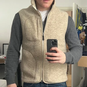 Duncan Wool Fleece Väst - J.Lindeberg - SÄLJER ELLER SÖKER BYTE, till en stilren beige väst i ull med dragkedja framtill. Perfekt för lager-på-lager under kyliga dagar eller bara som skönt tillägg till den outfit över en stickad tröja. Storlek M - Knappt använd och inga defekter. Nypris 2400:- 