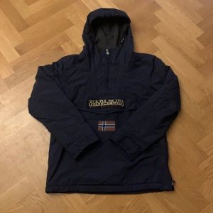 Mörkblå anorak från Napapijri - Säljer en mörkblå anorak från Napapijri med huva och dragkedja framtill. Jackan har en stor ficka på magen med Napapijri-logga och en norsk flagga. Perfekt för kyliga dagar. Använd ett fåtal gånger. Riktigt snygg och skön. Priset kan sänkas.