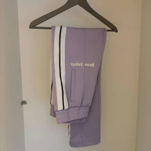 Lila mjukisbyxor från Palm Angels - Snygga lila mjukisbyxor från Palm Angels med vita och svarta ränder längs sidorna. Byxorna har elastiska muddar vid anklarna och en bekväm passform. Perfekta för en avslappnad stil.