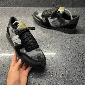 Valentino Rockrunner  - Snygga Valentino Rockrunner sneakers i svart och grått med ett coolt camouflage-mönster. Skorna har snörning och en unik design med nitar på hälen. Perfekta för att ge din outfit en edgy touch.