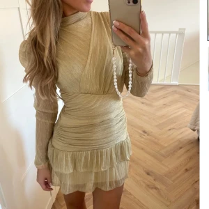 Guldglittrig klänning med volangdetaljer - Snygg guldglittrig klänning med långa ärmar och volangdetaljer. Den har en tight passform och en kort längd som ger en elegant look. Den är slutsåld på hemsidan! Passar mig som är vanligtvis xs och är 164cm lång, men storleken är S/M så passar dom flesta! Använd 2 gånger🤍den kostar 470kr + frakten som är 300kr på hemsidan! Så 770kr nypris💛