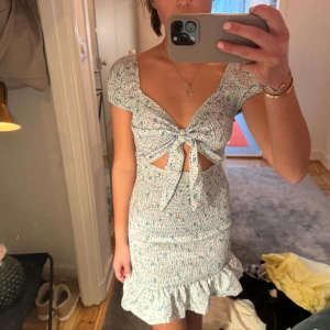 Blommig klänning med volangdetaljer -  blommig klänning i ljusa toner med volangdetaljer vid fållen. Klänningen har en knytning framtill och korta ärmar. Sitter så fint