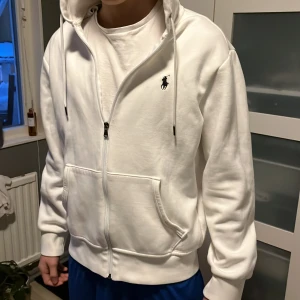 Vit hoodie från Ralph Lauren - Säljer en stilren vit hoodie från Ralph Lauren med dragkedja och justerbar luva. Den har en broderad logotyp på bröstet och praktiska fickor framtill. Perfekt för en avslappnad look. Nu pris 2295 jag säljer för 1299, använd väldigt få gånger. 