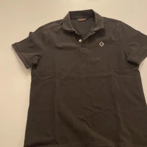 Svart pikétröja från Morris - Snygg grå/grön pikétröja från Morris med korta ärmar och en klassisk krage. Tröjan har en liten broderad logotyp på bröstet och knappar framtill. Perfekt för en stilren look. Grön/grå piké.