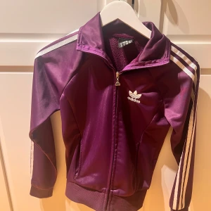 Lila träningsjacka från Adidas - Snygg lila träningsjacka från Adidas med klassiska vita ränder på ärmarna. Jackan har dragkedja och en broderad logga på bröstet. Sitter svin snyggt och lite croppad 