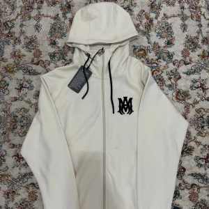 Hoodie från Amiri - Säljer en stilren hoodie från Amiri med svart logotyp på bröstet och ryggen. Hoodien har en dragkedja framtill och en justerbar huva med svarta snören. Perfekt för en avslappnad look.