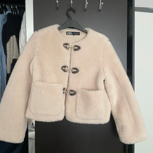 Beige teddyjacka från Zara - Skit snygg och mysig beige teddyjacka från Zara med dekorativa guldiga spännen framtill. Jackan har långa ärmar och stora fickor, perfekt för kyliga dagar. Den fluffiga texturen ger en trendig look. 