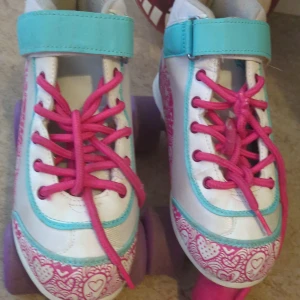 Färgglada rullskridskor med hjärtmönster - Säljer ett par supercoola rullskridskor i vitt med turkosa och rosa detaljer. De har rosa snörning och ett gulligt hjärtmönster runt tårna och sidorna. Perfekta för att rulla runt i parken eller på gatan!