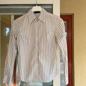 Randig skjorta från Peak Performance - Säljer en  randig skjorta från Peak Performance i storlek XS. Skjortan har en klassisk krage och långa ärmar. Den är vit med bruna och blå ränder, vilket ger den en stilren look. Perfekt för både vardag och lite mer uppklädda tillfällen. 