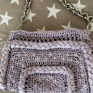 Ljuslila stickad axelväska med kedja - Säljer en superfin ljuslila stickad axelväska med en cool kedja som axelrem. Väskan har en dragkedja upptill för säker förvaring. Perfekt för att ge din outfit en unik touch!