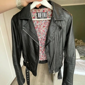 Svart skinnjacka från Jofama by Kenza - Svart skinnjacka i felfritt skick från Jofama by Kenza i storlek 34. Bikerstil med dragkedja och bälte i midjan. Nitar, dragkedja vid ärmslut, blommigt innerfoder. 