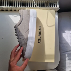 Axel Arigato skor - Säljer ett par snygga beige sneakers från Axel Arigato. De är gjorda i mocka och har en stilren design med vita sulor. Perfekta för både vanliga och finare tillfällen! Skorna har snörning och är i nyskick.