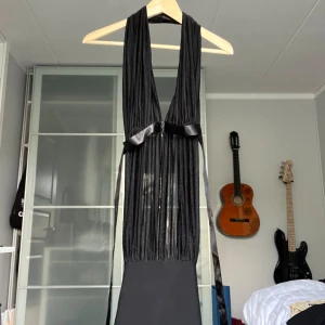 Svart klänning med öppen rygg och knytband - Säljer en elegant svart topp med en stilren design. Den har en öppen rygg och knytband i midjan. Toppen är halterneck och har en djup v-ringning framtill. Perfekt för festliga tillfällen!