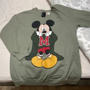 Grön tröja med Musse Pigg från H&M - Säljer en superfin grön tröja från H&M med en stor Musse Pigg-tryck på framsidan. Tröjan är långärmad och perfekt för en avslappnad stil. Den är i bra skick och passar perfekt för höst och vår. Disney-fans kommer älska denna!