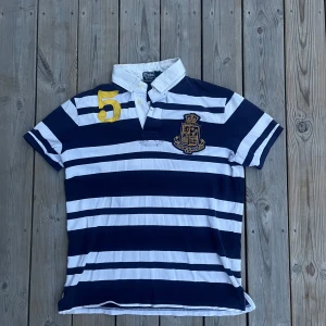 Randig pikétröja från Polo Ralph Lauren - Polo  från Polo Ralph Lauren i marinblått, embroidery och logga samt siffra på ryggen.. Sitter mer som M, men står L.