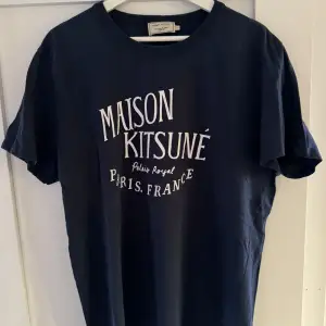 Säljer en snygg mörkblå t-shirt från Maison Kitsuné med tryck i vitt på framsidan. Den är kortärmad och perfekt för en avslappnad stil. T-shirten är i bra skick och passar både till vardags och fest.