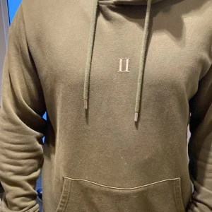 Olivgrön hoodie från Les Deux - Säljer en snygg olivgrön hoodie från Les Deux. Den är i bra skick och har en stilren design med en liten logga på bröstet. Perfekt för höst och vinter med sin mysiga känsla. Den har en klassisk passform med långa ärmar och en praktisk ficka framtill. Perfekt för både vardag och chill!