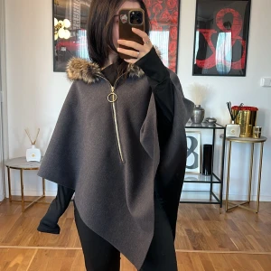 Grå poncho med fuskpäls - Säljer en superfin grå poncho med en mysig fuskpälsdetalj runt huvan. Den har en snygg dragkedja framtill med en cool ringdetalj. Perfekt för höst och vinter när du vill vara både varm och stilren. Passar perfekt över en långärmad tröja för extra värme.
