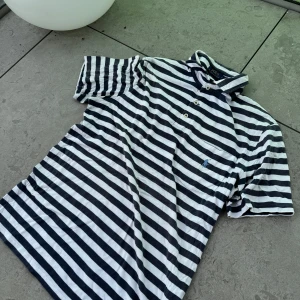 Randig pikétröja från Ralph Lauren - Säljer en klassisk randig pikétröja från Ralph Lauren i storlek M. Tröjan har en stilren design med vita och blå ränder och korta ärmar. Perfekt för en avslappnad sommarstil. Den är i bra skick och redo för nya äventyr!