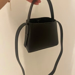 Svart axelväska med dragkedja - Snygg svart axelväska i skinn med stilren design. Väskan har en praktisk dragkedja upptill och en justerbar axelrem. Perfekt för att bära dina nödvändigheter med stil.