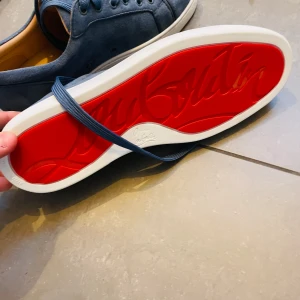 Blå sneakers med röd sula - Snygga blå sneakers med den ikoniska röda sulan. Skorna har snörning och en stilren design som passar perfekt för en trendig look. Den blå färgen kombineras med en vit sula för en fräsch kontrast. Shoebox och allt annat og och autenticitet kort finns med. Pris kan diskuteras
