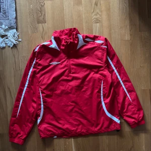 Röd vindjacka från Umbro - Säljer en röd vindjacka från Umbro med vita detaljer. Jackan har en halv dragkedja framtill och långa ärmar. Perfekt för sportiga aktiviteter eller som en lätt ytterjacka. 🏃‍♂️