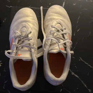 Vita Adidas Copa fotbollsskor - Säljer ett par vita Adidas Copa fotbollsskor med orange detaljer och dobbar. Skorna har snörning och är perfekta för fotbollsspel på gräs. De har en stilren design med Adidas klassiska ränder i orange på sidan.