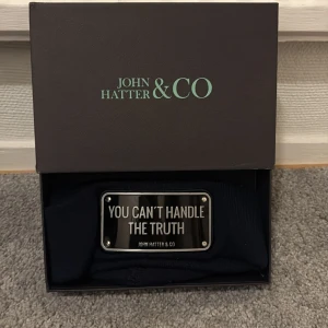 Svart mössa från John Hatter & Co - Snygg svart mössa från John Hatter & Co med en metallplatta framtill där det står 'YOU CAN'T HANDLE THE TRUTH'. Perfekt för att ge en cool touch till din outfit. Den är lite sönder med. 