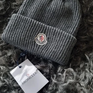 Grå ribbad mössa från Moncler - Snygg grå ribbad mössa från Moncler med broderad logotyp framtill. Perfekt för kyliga dagar och ger en stilren look. Mössan är tillverkad i ett mjukt och bekvämt material.