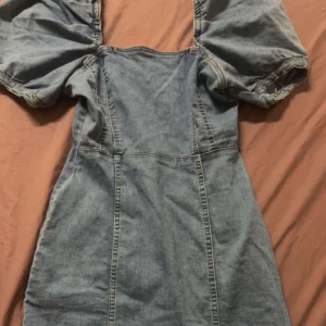 Jeansklänning från H&M Divided - Trendig jeansklänning från H&M Divided med puffiga kortärmar och en figurnära passform. Perfekt för en avslappnad stil med även retro. Klänningen har en klassisk blå jeansfärg och är tillverkad i ett bekvämt material.