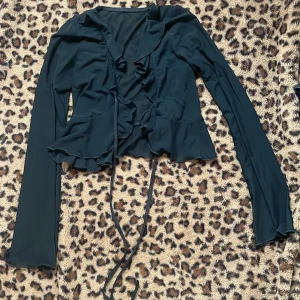 Svart långärmad blus med volangdetaljer i mesh - Elegant svart blus med långa ärmar och volangdetaljer framtill. Blusen har en knytning i midjan och en lätt croppad passform. Perfekt för en stilfull look.