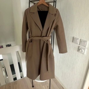 Beige kappa från Amisu - Snygg beige kappa från Amisu i storlek S. Den har en klassisk design med knytbälte i midjan och långa ärmar. Perfekt för höst och vår, denna kappa ger en stilren look.