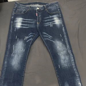 Blå jeans med slitningar dsq*eared - Snygga blå jeans med coola slitningar och en klassisk femficksdesign. De har en knapp och dragkedja framtill samt en röd etikett vid fickan. Perfekta för en avslappnad stil.