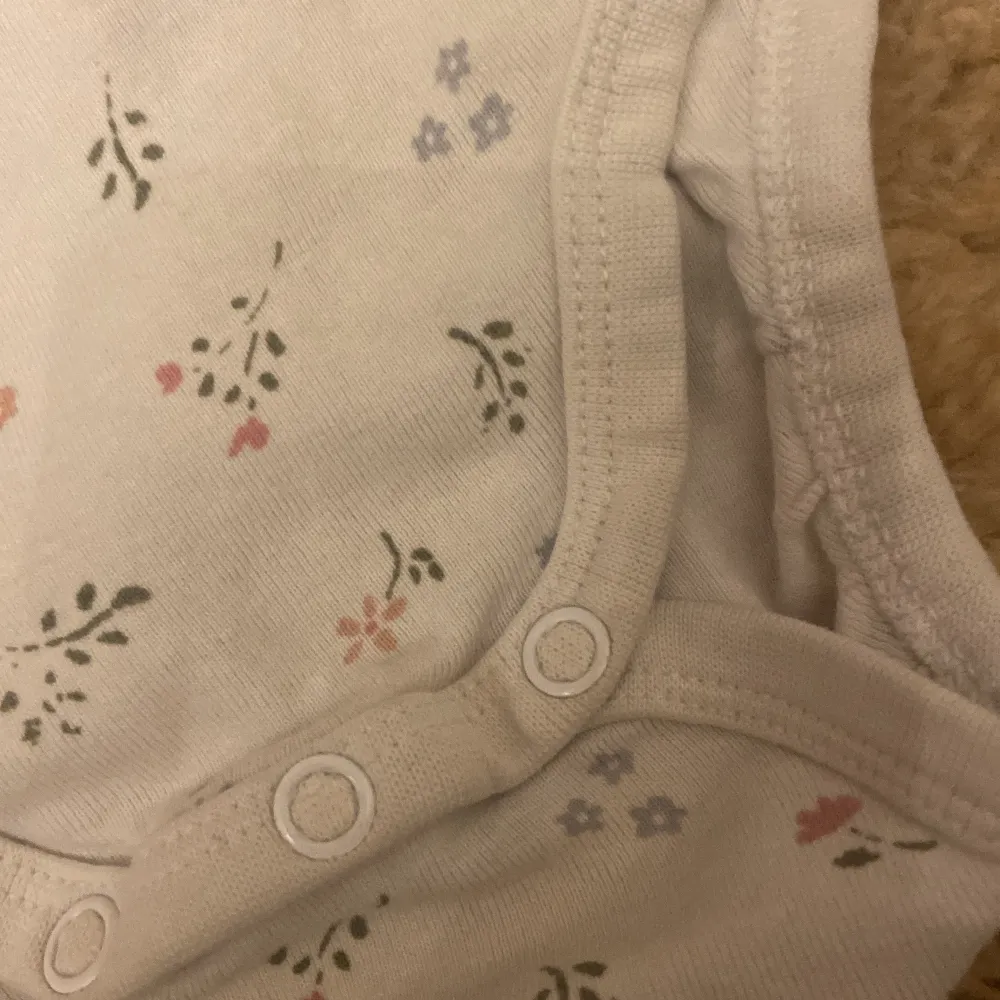 Söt vit body från H&M i ekologisk bomull med små blommor i rosa, blått och grönt. Den har en liten rosett vid halsen och praktiska tryckknappar i grenen. Perfekt för de små! 🌸. Neuletakit & Villapaidat.