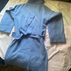 Blå stickad klänning med bälte - Mysig blå stickad klänning med polokrage och trekvartsärmar. Den har ett matchande bälte i midjan för en snygg passform. Perfekt för kyligare dagar.