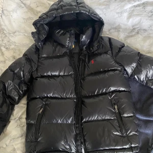 Ralph Lauren Jacka - Snygg svart pufferjacka med glansig finish och huva. Jackan har dragkedja framtill och två fickor med dragkedjor. Perfekt för kyliga dagar.