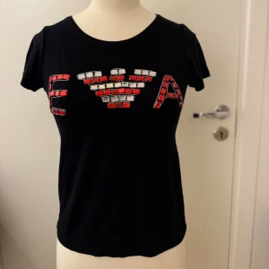 Emporio Armani Svart t-shirt  - Snygg svart t-shirt från Emporio Armani med korta ärmar. XS /S Framsidan pryds av ett mönster med röda och vita stenar som bildar bokstäverna E/A. Hämtas på Hägerstensåsen eller ök 🖤