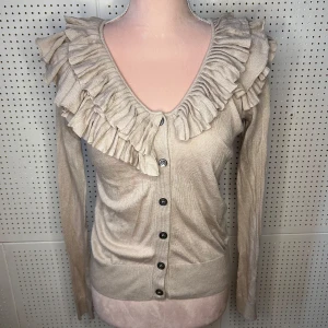 Beige kofta med volangdetaljer - Säljer en elegant beige kofta med volangdetaljer runt halsringningen. Koftan har långa ärmar och knäpps med knappar framtill. Perfekt för en stilren look.