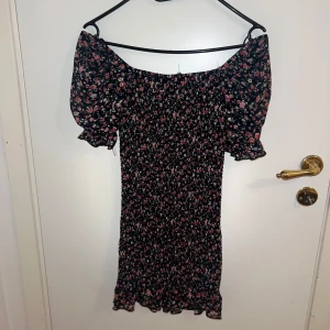 Blommig offshoulder klänning - Söt blommig klänning med offshoulder-design och korta ärmar. Den har ett elastiskt band vid axlarna och volangdetaljer vid ärmslut och nederkant. Perfekt för sommarens varma dagar.