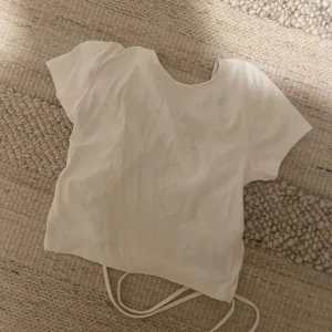 Vit kortärmad topp från Gina Tricot - Säljer en stilren vit topp från Gina Tricot. Den har korta ärmar och en rund halsringning. Toppen är croppad och har en knytning i ryggen för en snygg detalj. Perfekt för varma dagar!