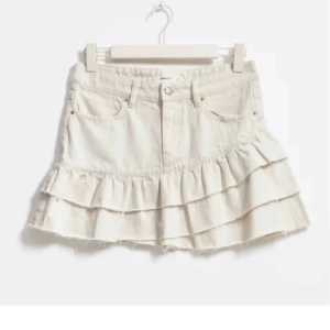 Beige jeanskjol med volanger - Trendig beige jeanskjol med volanger och råa kanter. Kjolen har en kort längd och klassisk femficksdesign. Perfekt för en avslappnad och stilren look.