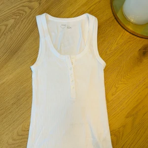 Vit ribbad linnetopp från American eagle - Säljer en vit ribbad linnetopp från ae med knappar framtill. Perfekt för varma dagar och enkel att matcha med olika outfits. Toppen har en klassisk design med rund halsringning.