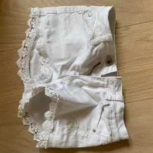 Vita shorts med spetsdetaljer - Snygga vita shorts med spetsdetaljer längs kanterna. Perfekta för sommarens varma dagar. De har en knapp och dragkedja framtill samt klassiska fickor.