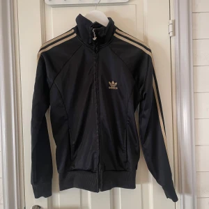 Svart träningströja - Snygg svart träningströja från Adidas med klassiska tre ränder i guld längs ärmarna. Fortfarande jättebra skick. Kan vara vintage men finns inga lappar kvar förutom den på bilden. Fick den av en familjekompis så ingen aning när den är ifrån. Använder inte. Om man vi ser hur den ser ut på är det bara att fråga😊