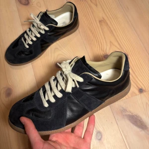 Maison Margiela gats - Snygga svarta sneakers från Maison Margiela med en stilren design. Skorna har en kombination av mocka och skinn, vilket ger en lyxig känsla. De har vita snören och en brun sula som ger en fin kontrast. Perfekta för en trendig look.