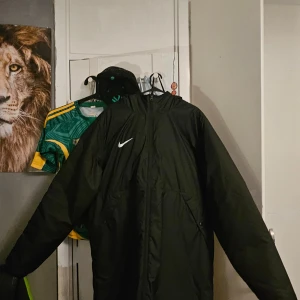 Svart lång jacka från Nike - Säljer en svart lång jacka från Nike med luva och dragkedja. Jackan har en loose fit och är perfekt för kyligare dagar. Den har en stor logga på baksidan och är tillverkad i ett slitstarkt material.