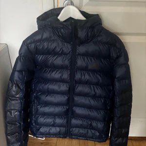 Mörkblå dunjacka shine NY st L från Adidas - Säljer en snygg mörkblå dunjacka från Adidas med huva och dragkedja. Jackan har de klassiska tre ränderna på ärmarna och är perfekt för kyligare väder. Adidas shine jacka st L. Blå (sista bild visar bäst färgen). Sidfickor med dragkedja. Adidas-logga på bröstet och 3-Stripes längs ärmarna. Kan maskintvättas. Blev fel storlek så aldrig använd.  Nypris 1200 kr. 